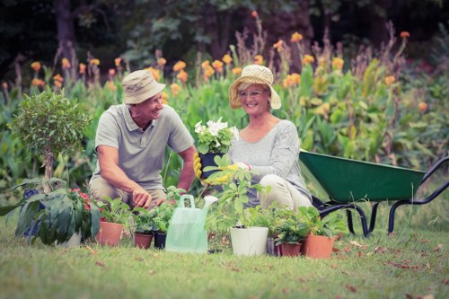 A cheerful elderly couple in a garden, wearing wide-brimmed hats and gardening gloves, are seated on the grass surrounded by an assortment of potted plants and gardening tools. The woman is smiling while holding a white flowering plant, and the man is leaning towards her, appearing engaged in conversation. Behind them, there are vibrant purple, orange, and yellow flowers blooming in a well-maintained flower bed with lush green foliage. To their side, a green garden wheelbarrow is partially visible, and a watering can sits on the ground among the plants. The scene is set outdoors on a sunny day, with a backdrop of trees and shrubbery, creating a peaceful, natural environment that showcases a well-tended garden. This scene reflects typical gardening activities and outdoor enjoyment, aligning with landscaping and garden maintenance services such as those offered by Landscapers Putney in the Putney area, south-west London, with the setting suggesting a local suburban garden environment.