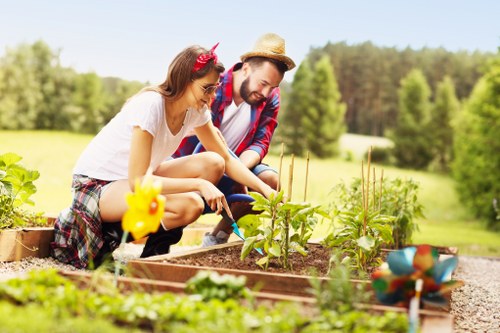A woman and a man are working together in a well-maintained backyard garden, tending to a raised bed containing young plants and seedlings. The garden features a grassy lawn area with lush, green grass in the foreground, bordered by wooden edging. The background displays a neatly trimmed hedge and tall, mature trees under a clear, sunny sky, suggesting a bright summer day. The garden environment includes a pathway with gravel or stone, surrounding the planting beds, which contain rich, dark soil. There are garden tools and decorative items, such as a yellow watering can and a small ceramic animal figurine, visible near the planting area. The scene offers a natural, inviting outdoor space designed for gardening and outdoor leisure, and it subtly emphasizes the services offered by Landscapers Putney for garden maintenance and landscaping in the local area near Putney, South West London, enhancing the site’s local SEO relevance.