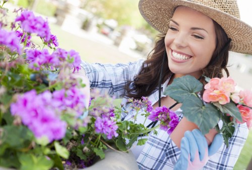 A woman in a wide-brimmed straw hat and light blue gardening gloves is shown smiling as she tends to a variety of vibrant flowering plants in a garden. In the foreground, there are clusters of purple, pink, and orange flowers, with green foliage providing a lush backdrop. The garden features a neatly maintained lawn with detailed edging along flower beds, and visible pathways or paving stones can be seen in the background, hinting at a well-organised outdoor space typical of suburban gardens in the Putney area. The scene appears to be during a bright, sunny day, with natural light illuminating the garden elements and the gardener's cheerful expression. This image reflects professional gardening care, such as planting and tending to flower beds, supporting outdoor landscaping services offered by companies like Landscapers Putney, which specialise in garden maintenance and enhancement within the local area.