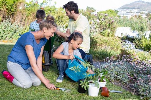 A family garden scene in a landscaped outdoor space, featuring a well-maintained lawn bordered by flower beds with a variety of flowering plants and shrubs in natural green, purple, and white tones. In the foreground, a woman wearing a blue top is kneeling on the grass, planting or tending to a small plant with a garden fork nearby. A young girl, dressed in casual clothes, is sitting on the ground with a blue backpack open, handling a garden tool or plant, with a white watering can and a small terracotta pot beside her. In the background, a man with dark hair, wearing a white shirt, stands with another child and appears to be engaged in gardening activities. The setting is in natural daylight with slight cloud cover, suggesting mild weather. The area shows clear, evenly cut grass, neat flower beds, and a landscaped environment that aligns with professional gardening services by Landscapers Putney, suitable for yard upgrades or patio repair projects in SW15 or SW14 postal districts near Putney.