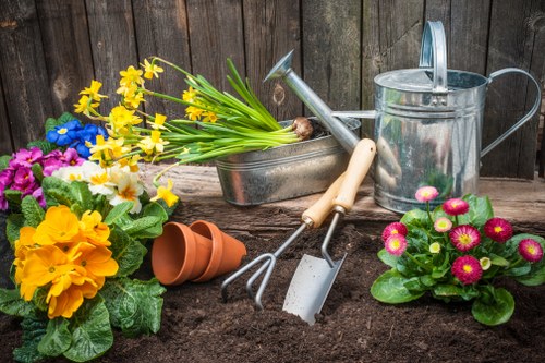 A garden scene featuring a freshly prepared planting bed with rich, dark soil, surrounded by a variety of colorful flowering plants including pink primulas and yellow primroses, set against a wooden fence backdrop. Gardening tools such as a trowel, hand fork, and a watering can are placed nearby, along with plant pots and a small watering can, indicating ongoing planting or garden maintenance work. The garden environment appears well-maintained with a mix of structured flower beds and natural plant arrangements, reflecting typical gardening activities in a residential outdoor space within Putney. The weather appears clear with natural daylight illuminating the scene, emphasizing the vibrant colors of the flowers and the tools used for caring for the garden. This detailed description supports gardening services including planting, soil preparation, and outdoor maintenance, relevant to local gardens in the Putney area.