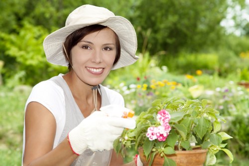 A woman in a white sun hat and gardening gloves is smiling while tending to a potted pink-flowered plant in a lush, green garden. The garden features a well-maintained lawn with dense grass, bordered by a variety of shrubs and flowering plants. Behind her, there are taller trees and a vibrant array of colorful flowers, creating a lively outdoor setting with natural sunlight illuminating the scene. The overall environment appears to be a landscaped backyard garden, typical of outdoor spaces in Putney, with an emphasis on healthy plant growth and garden care. The scene highlights the importance of regular gardening activities, consistent with professional lawn and garden maintenance services provided by companies like Landscapers Putney, reflecting a vibrant and thoughtfully designed outdoor area suitable for local gardens in the SW15 postcode area.
