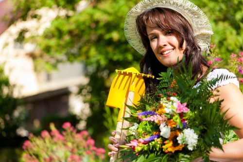 A woman with shoulder-length brown hair wearing a wide-brimmed straw hat is standing in a well-maintained garden, holding a large bouquet of mixed flowers and greenery. Behind her, there is a lush green hedge and a lawn area with neatly trimmed grass, interspersed with flowering plants and shrubs. The garden appears to be part of a residential outdoor space, featuring a combination of planted borders and open grassy sections, with sunlight filtering through trees indicating clear weather. The scene emphasizes natural garden elements typical of a landscaped yard, including soil beds, flowering bushes, and a tidy appearance maintained by professional gardening services. This setting, typical of residential gardens in Putney, highlights the type of outdoor spaces landscaped and cared for by companies like Landscapers Putney, which specializes in garden care and landscaping projects.