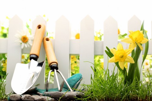 A close-up view of a small garden space with a white picket fence in the background, featuring a patch of vibrant green grass and a flower bed with bright yellow daffodils and white daisies. In the foreground, garden tools including a trowel and a small hand rake with wooden handles rest on the soil, indicating recent or ongoing gardening work. The garden area is well-lit by natural sunlight, highlighting the lively colours and healthy plant growth typical of a well-maintained outdoor space in Putney, London. The scene reflects elements of garden landscaping and lawn care, emphasizing a neat and inviting outdoor environment that a local gardening service such as Landscapers Putney would support, with a focus on planting, lawn maintenance, and garden tidying efforts typical of the area.