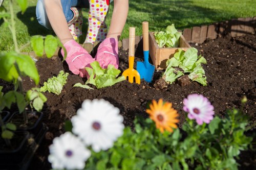 A person wearing pink gardening gloves is tending to a garden bed in a backyard, using small gardening tools such as a yellow hand rake, a blue trowel, and a wooden plant marker. The garden contains a variety of leafy green vegetables, including lettuce, planted in dark, freshly turned soil. In the foreground, there are colorful flowering plants with white, purple, and orange blooms, adding vibrant contrast to the scene. The background features a well-maintained grassy lawn with lush green grass, indicating a healthy outdoor space possibly located in Putney, London. The setting appears to be a bright, sunny day, enhancing the natural colors of the garden and promoting outdoor gardening activities. This image reflects a typical urban garden layout with designated vegetable beds, flower borders, and gardening tools, aligning with professional gardening services offered by Landscapers Putney, including lawn and garden bed maintenance.