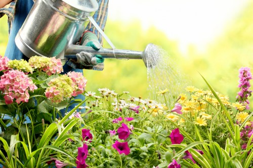 A gardener's hand, wearing a garden glove, is holding a silver watering can and watering a diverse garden bed featuring pink, yellow, and purple flowers. The garden includes lush green foliage, grass, and flowering plants arranged in a mixed bed setting. The scene shows natural daylight with soft background greenery, suggesting an outdoor garden space in early summer or late spring. The image highlights outdoor gardening activities typical of professional lawn and garden maintenance services provided by Landscapers Putney, with a focus on plant care and floral cultivation in a residential or landscaped garden in Putney, London.