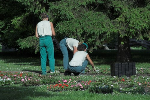 The image depicts three individuals working in a lush, well-maintained garden with dense green trees and a grassy lawn. Two individuals are actively planting or tending to a flower bed bordered by a variety of small, colorful flowers in shades of pink, red, and white. One person is kneeling down, closely working on the plants, while another is bending over, possibly assisting in planting or weeding. The third individual is standing nearby, observing or preparing to help. The garden scene features a mix of soft grass, flower beds, and mature trees providing shade, with a natural and orderly outdoor environment typical of residential gardens in Putney or surrounding southwest London areas. The lighting suggests a clear day with natural sunlight, highlighting the vibrant green foliage and blooming flowers. This scene reflects professional gardening or lawn care activities, consistent with services offered by companies like Landscapers Putney, supporting garden restoration, planting, and outdoor maintenance.