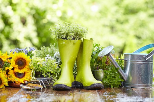 A garden scene featuring a wooden outdoor table with a pair of bright green rubber gardening boots filled with white and light green flowering plants, positioned next to a collection of sunflower and mixed flower bouquets. To the right, a metal watering can with a blue handle and a garden trowel resting inside are visible, all set against a blurred background of lush green foliage and sunlight filtering through trees. The scene highlights outdoor gardening tools and decorative plant arrangements, typical of a well-maintained garden space in Putney, suitable for professional landscaping or gardening services aimed at enhancing outdoor environments.