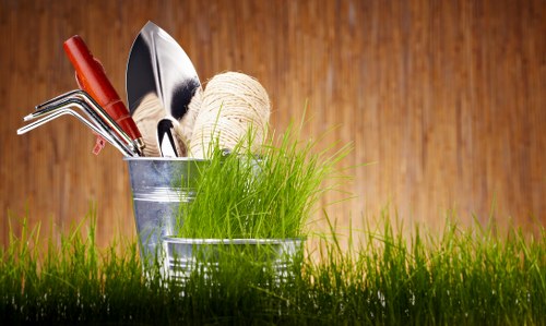A close-up of gardening tools and supplies arranged outdoors on a grass surface, with a wooden fence in the background. The tools include a gardening fork with a red handle, a trowel, and a pair of pruning shears, all resting inside a small metal bucket. Next to it is a second metal container filled with bright green grass or young plants. The scene suggests preparation for garden planting or maintenance, with natural sunlight illuminating the vibrant green grass in the foreground and warm wooden tones behind, highlighting a typical outdoor garden setting in the UK, suitable for professional landscaping or lawn care activities by companies such as Landscapers Putney.