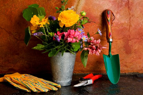 A close-up of a gardening workspace showing a large grey concrete planter filled with a colorful assortment of fresh flowers and green foliage, including yellow, pink, and purple blooms, positioned on a dark outdoor surface. To the right of the planter, there is a small green hand trowel with a multicoloured wooden handle, leaning against a reddish-brown textured wall, alongside a pair of red gardening shears with black handles. On the left, a pair of yellow and orange striped gardening gloves rests on the surface. The scene depicts an outdoor space suitable for planting and garden maintenance, with natural lighting highlighting the vibrant plant colors and textured surfaces, integrating elements typical of gardening services in the Putney area.
