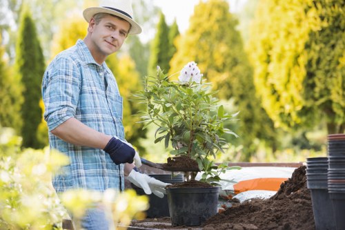 The image shows a man in a garden during a sunny day, wearing a light-colored fedora hat, a blue plaid shirt, and gardening gloves, tending to a potted flowering plant with pink and white blossoms. He is standing at a garden table with a partially dug soil area nearby, surrounded by various gardening supplies such as empty plant pots and gardening tools. The background features lush green trees and shrubs, creating a vibrant and well-maintained outdoor space typical of a suburban garden in Putney, London. The scene emphasizes outdoor gardening activities, highlighting the importance of plant care and landscape maintenance, and could relate to professional gardening and landscaping services offered by companies like Landscapers Putney. The natural lighting enhances the lush greenery and adds warmth to the scene, reflecting a peaceful, cultivated garden environment suitable for gardening services both for private gardens and communal outdoor spaces.