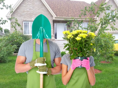 A person wearing a grey t-shirt, green gardening gloves, and a green apron, holding a teal garden shovel with a curved handle, stands in front of a well-maintained front garden. Another person, also wearing gardening gloves and a grey t-shirt, holds a large pink pot filled with bright yellow flowering plants, obscuring their face. The garden features a neatly trimmed lawn with dense green grass, a variety of small shrubs, and young trees with light green foliage. In the background, there is a brick house with white-framed windows and a grey roof, partially visible behind the plants. The scene appears to be on a bright but overcast day, with soft natural light illuminating the outdoor space. This image reflects typical gardening and landscaping activities in a suburban Putney garden, suitable for a service page about lawn and garden maintenance or plant care offered by Landscapers Putney.