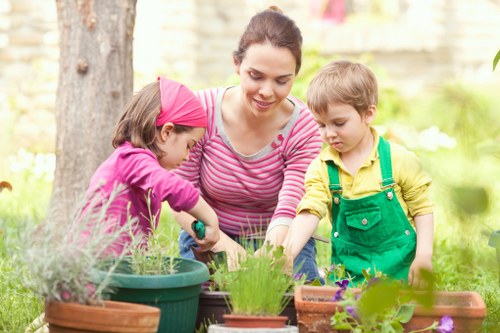 The image depicts a woman and two young children gardening outdoors in a well-maintained backyard, likely in Putney. The woman, dressed in a pink and white striped top, is smiling and overseeing the children's activity as they work together on a gardening project. The children, a girl wearing a pink headband and a purple jacket, and a boy in a yellow shirt with green overalls, are focused on planting in a variety of pots and garden beds. The garden features a green lawn in the foreground, surrounded by potted plants, flower beds, and a large mature tree providing shade in the background. The scene is illuminated by natural daylight, suggesting a clear, pleasant day, with lush greenery and some flowering plants visible. The setting showcases typical elements of a landscaped garden, including soil beds, container plants, and wooden garden furniture, reflecting a peaceful and vibrant outdoor space suitable for family gardening activities, which aligns with gardening and outdoor maintenance services provided by Landscapers Putney.